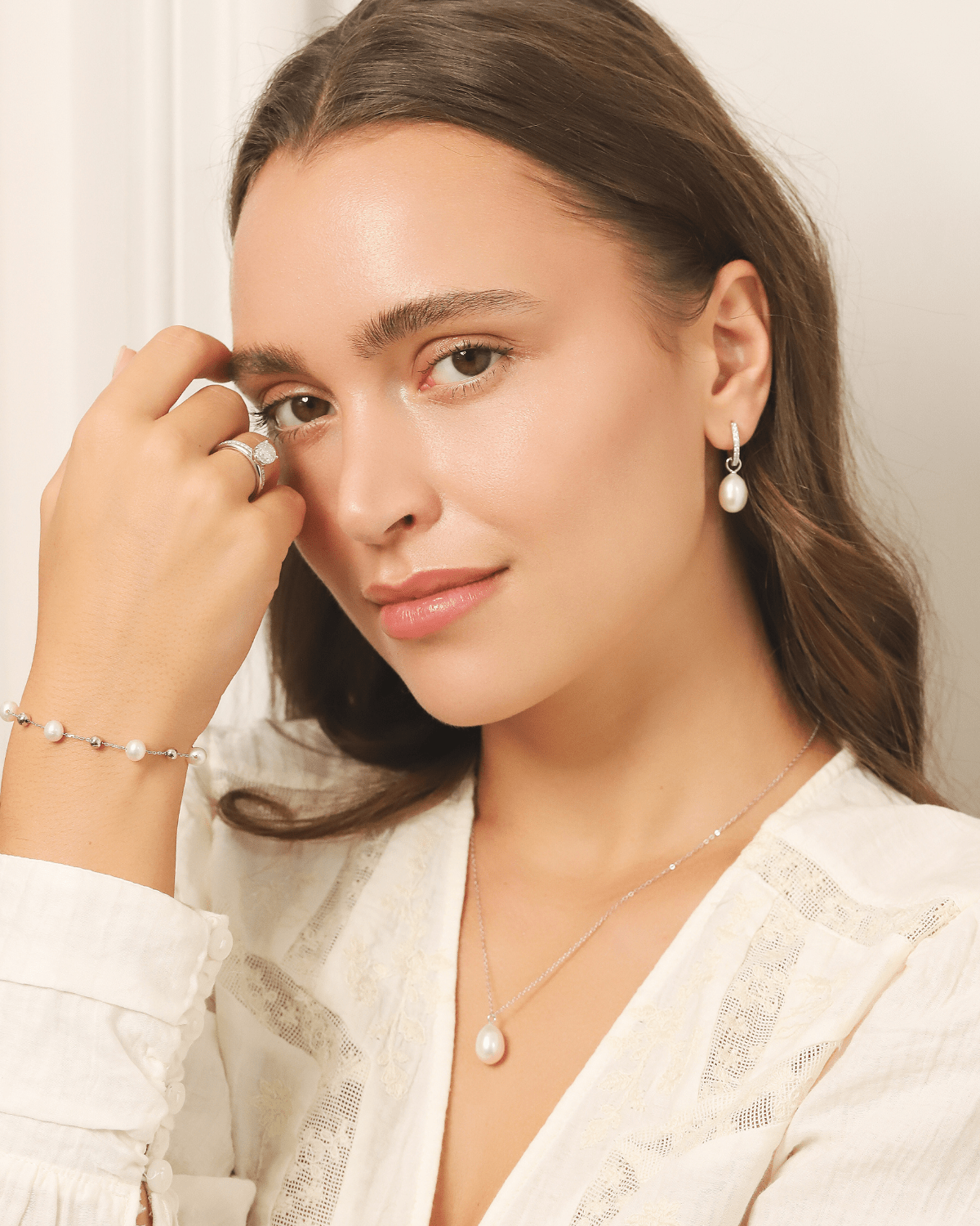 A woman wearing a white blouse, pearl earrings, and a delicate pearl necklace.