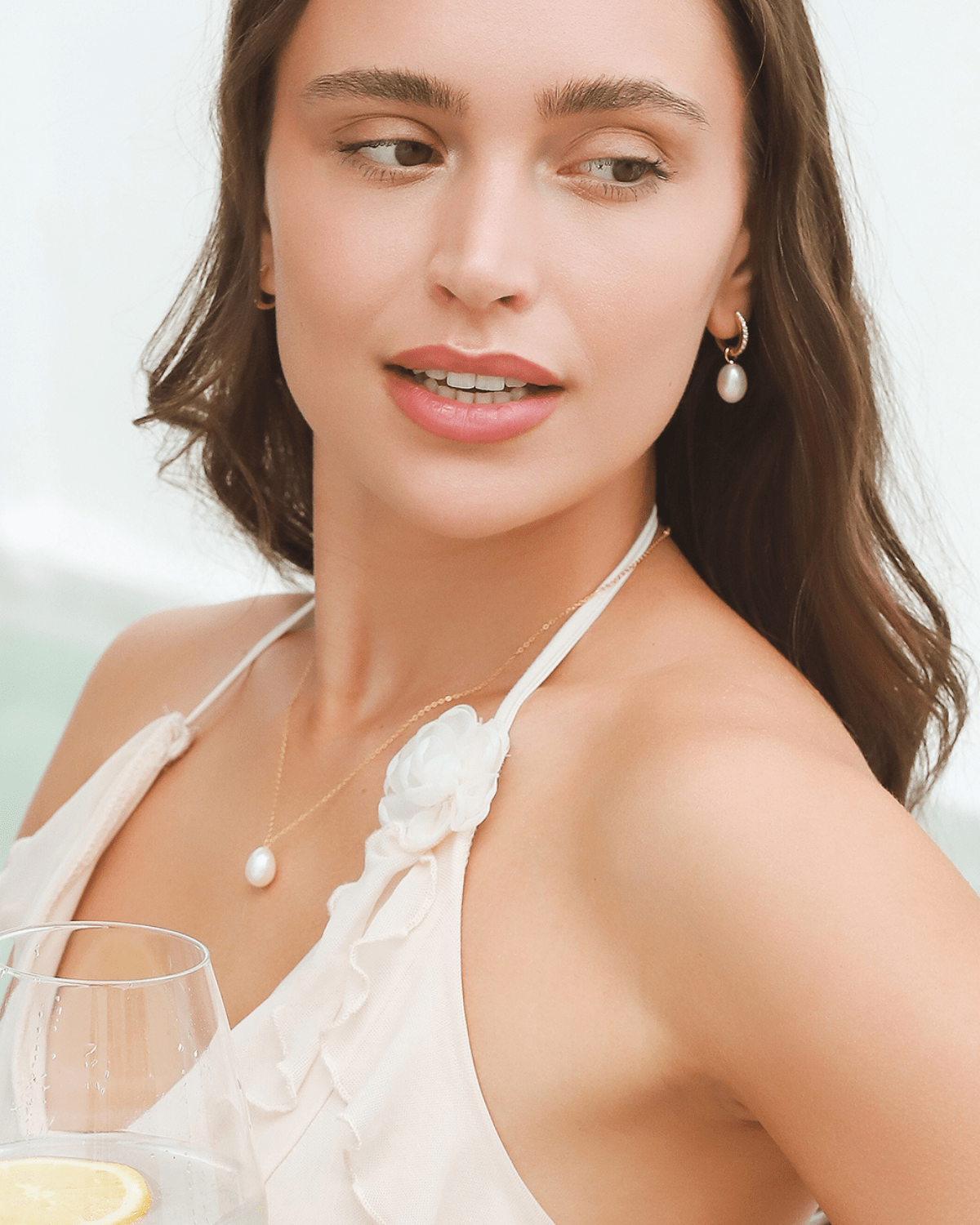 A woman wearing a pearl necklace and earrings, holding a drink and looking away.