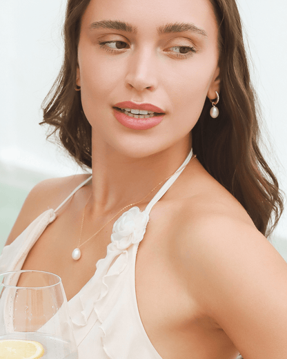 A woman wearing a pearl necklace and earrings, holding a drink and looking away.