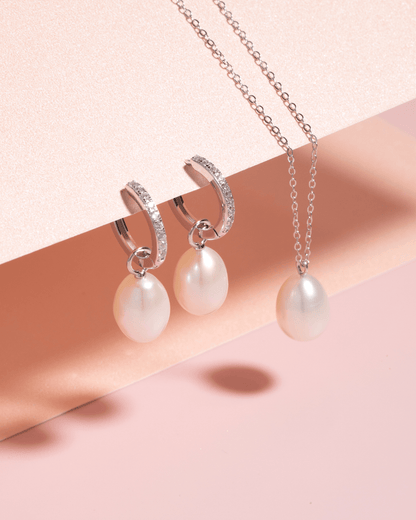 A pearl pendant necklace and pearl drop earrings displayed on a pink surface.