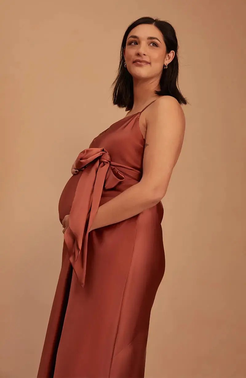Pregnant Woman wearing a terracotta brown bridesmaid dress with a bow detail on a beige background