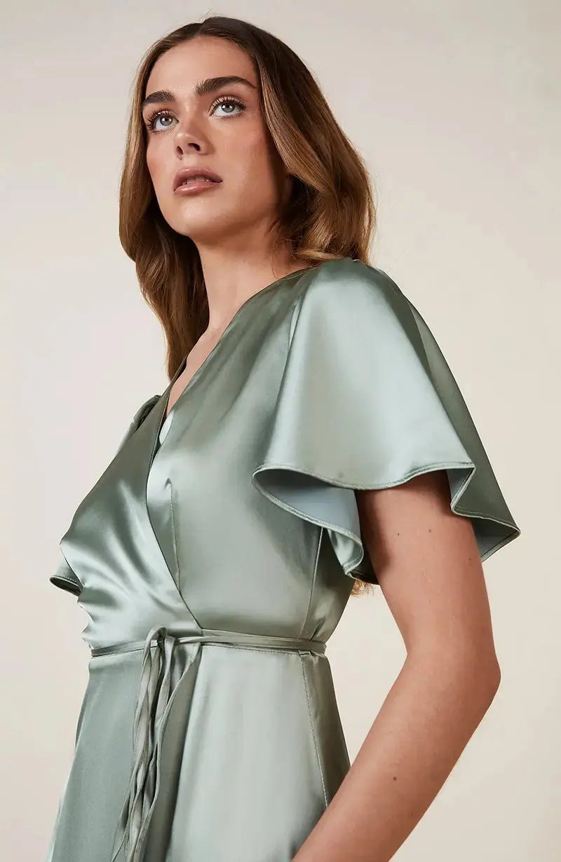 Woman wearing a light sage green satin Bridesmaid dress with ruffled sleeves on a plain background