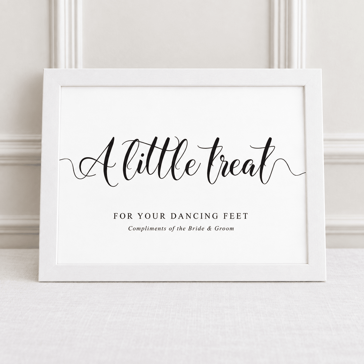 A white framed typographic wall print with black script reading “A little treat” and smaller text “FOR YOUR DANCING FEET / Compliments of the Bride & Groom,” leaning against a light wall.