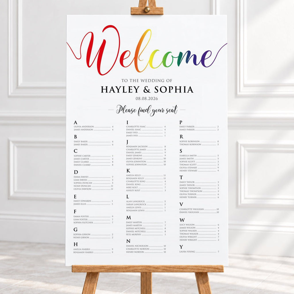 A vertical wedding welcome sign displayed on a wooden easel in a bright indoor setting. The sign has a white background with a large rainbow 'Welcome' script at the top and black seating chart text below.