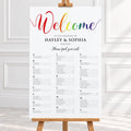 A vertical wedding welcome sign displayed on a wooden easel in a bright indoor setting. The sign has a white background with a large rainbow 'Welcome' script at the top and black seating chart text below.
