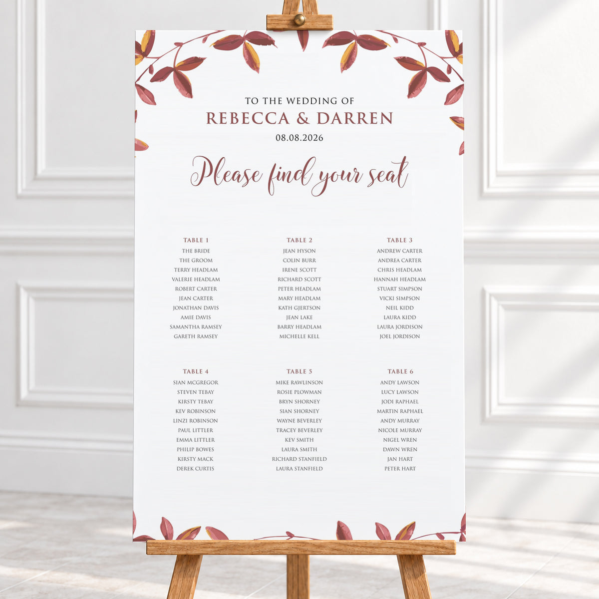 A wedding seating chart print with a white background, elegant brown and dusty rose typography, and a border of autumn leaves in muted red, brown, and gold tones with 6 wedding tables. The print is shown hanging from a wooden hanger on a light-colored wall above a white surface.