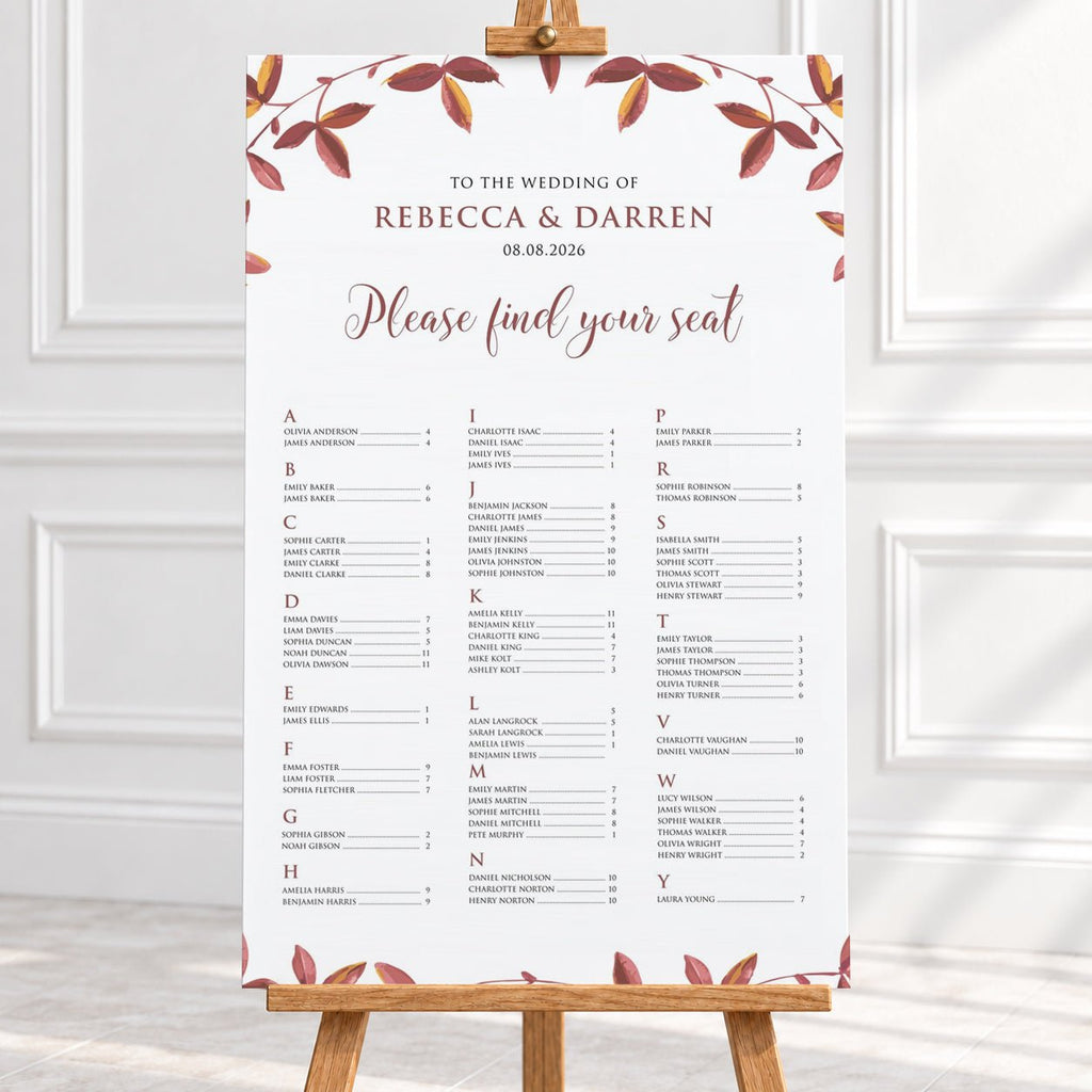A wedding seating chart printed on a large white board with elegant floral borders in muted autumn red and brown tones, displayed on a wooden easel in a bright, clean interior setting at a fall wedding.