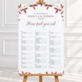 A wedding seating chart printed on a large white board with elegant floral borders in muted autumn red and brown tones, displayed on a wooden easel in a bright, clean interior setting at a fall wedding.