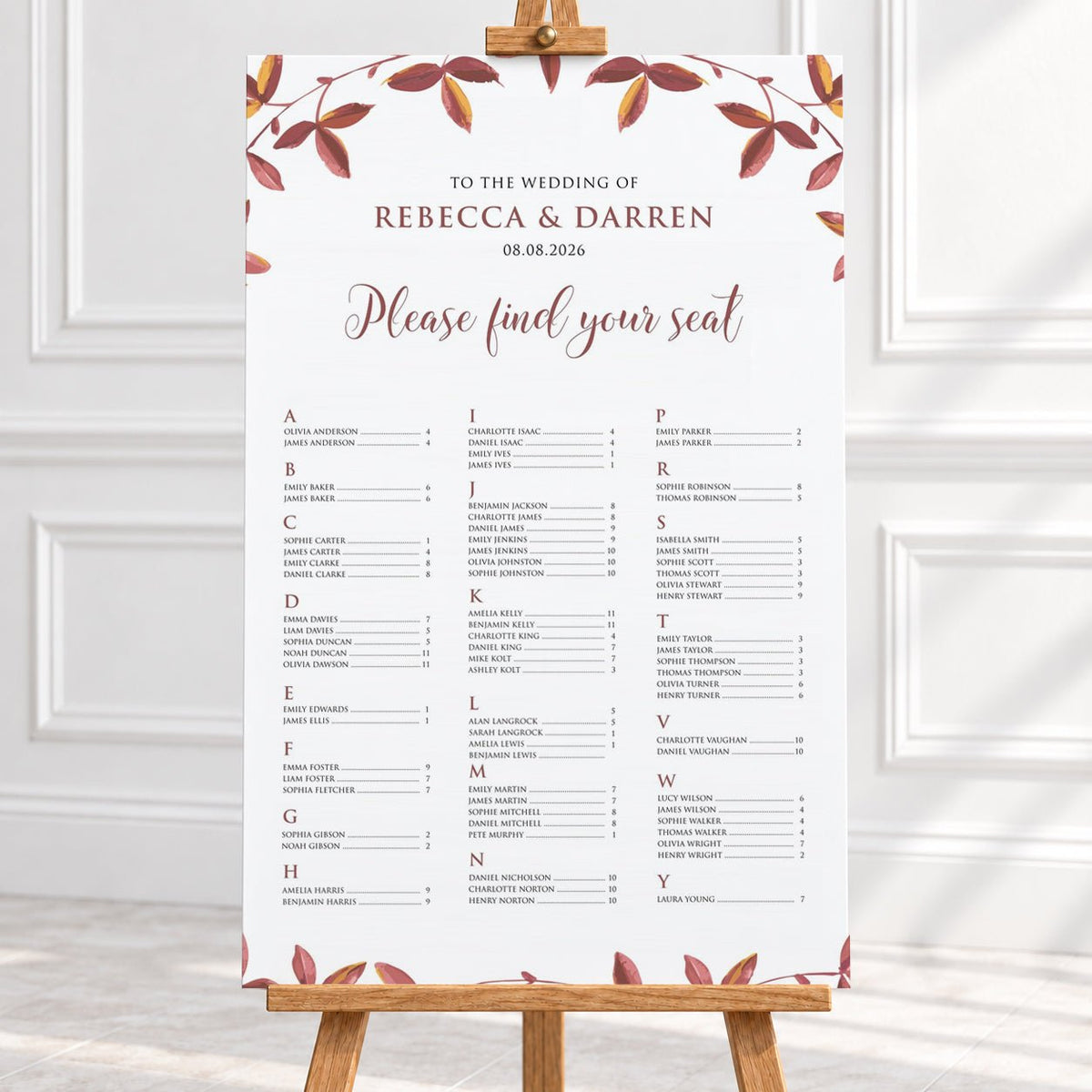 A wedding seating chart printed on a large white board with elegant floral borders in muted autumn red and brown tones, displayed on a wooden easel in a bright, clean interior setting at a fall wedding.