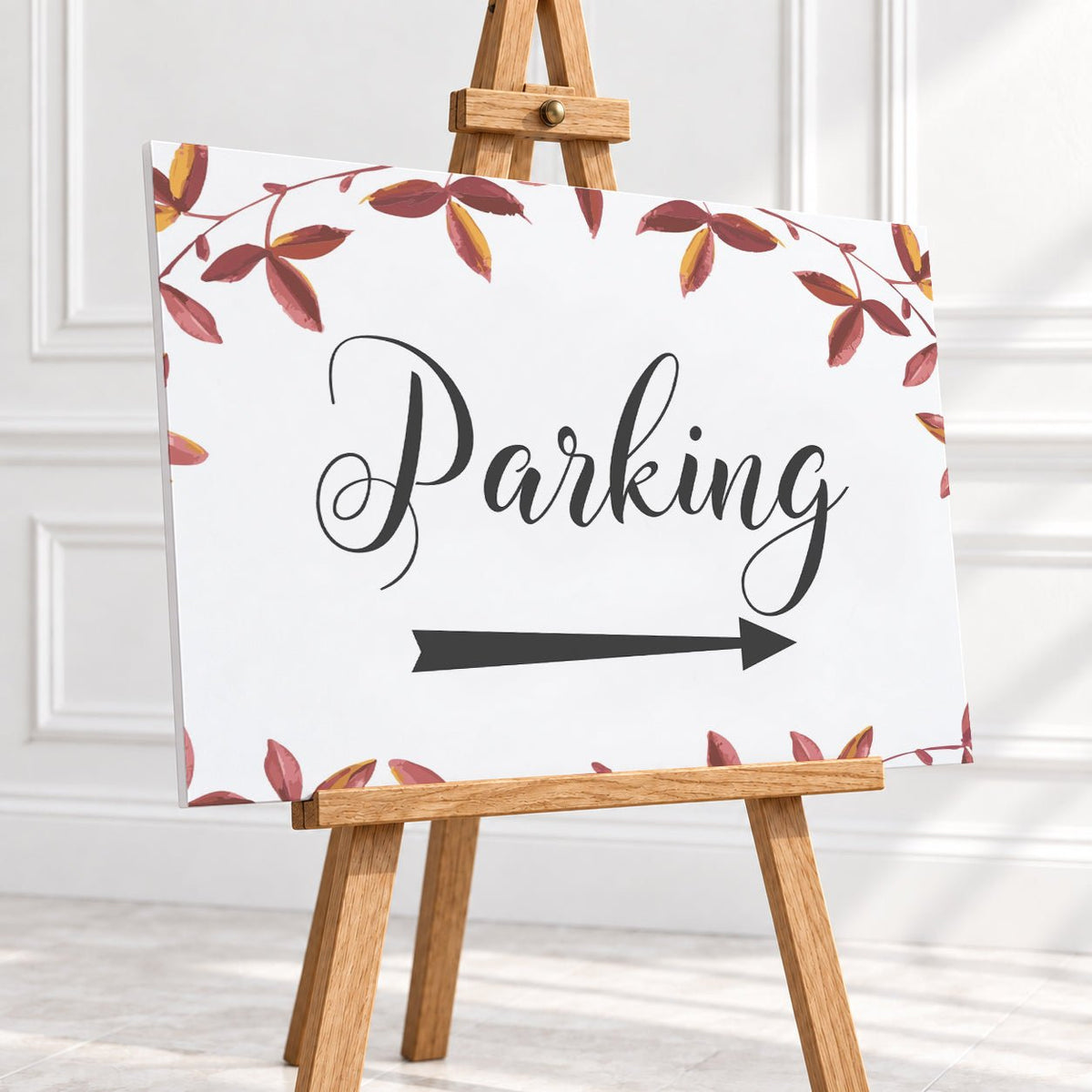 A rectangular white sign with the word 'Parking' in black script and a black right-pointing arrow, surrounded by a border of red and brown leaves. The sign is displayed on a small wooden easel in front of a white paneled wall.