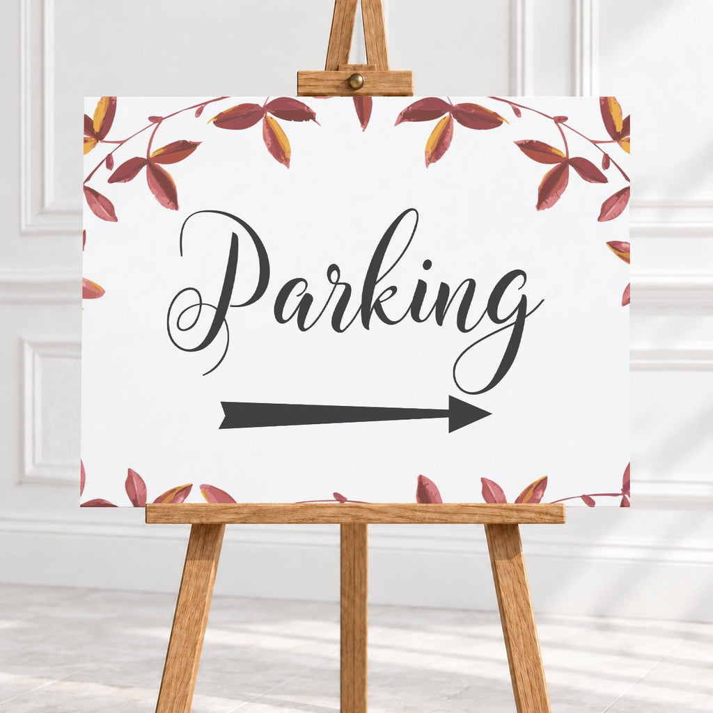 A vertical white sign with the word 'Parking' in black script, a black arrow pointing right, and a border of red and brown autumn leaves, displayed on a wooden easel in a bright indoor setting.