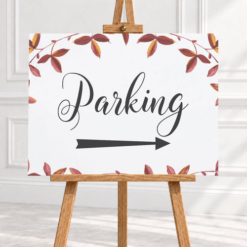A vertical white sign with the word 'Parking' in black script, a black arrow pointing right, and a border of red and brown autumn leaves, displayed on a wooden easel in a bright indoor setting.