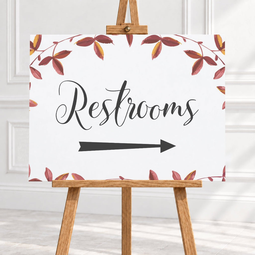 A restroom sign on a wooden easel, featuring the word 'Restrooms' in black script with a black arrow pointing right and a watercolor border of red and brown leaves on a white background.