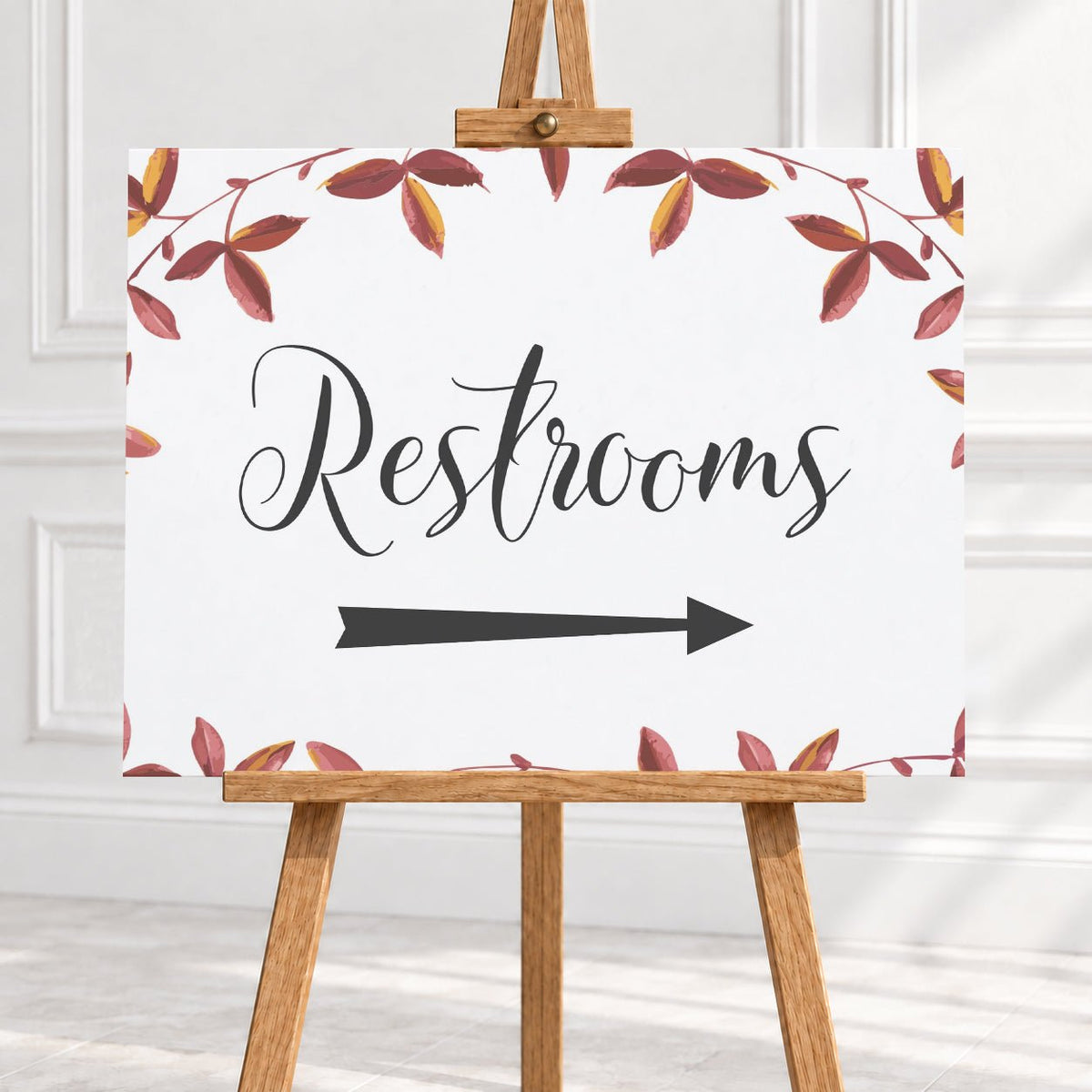 A restroom sign on a wooden easel, featuring the word 'Restrooms' in black script with a black arrow pointing right and a watercolor border of red and brown leaves on a white background.