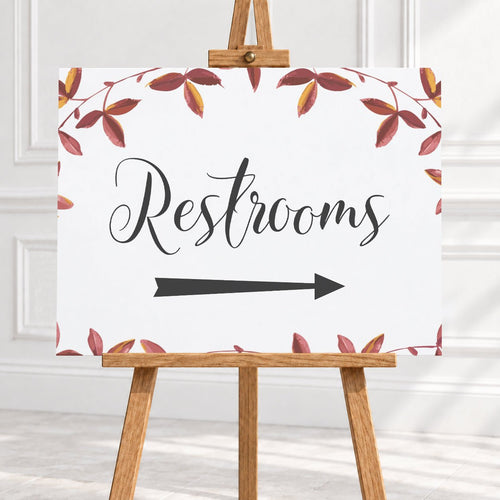 A restroom sign on a wooden easel, featuring the word 'Restrooms' in black script with a black arrow pointing right and a watercolor border of red and brown leaves on a white background.