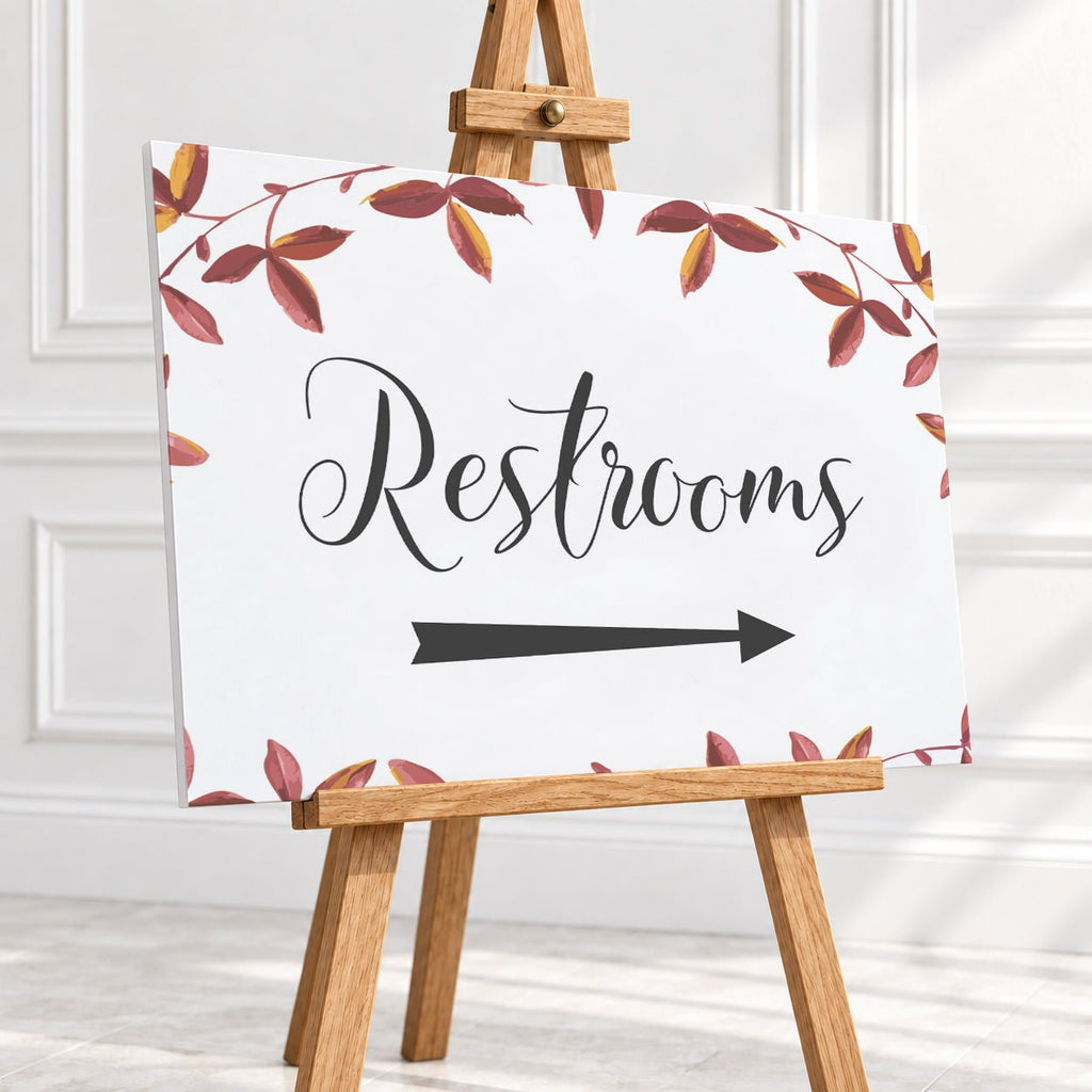 A rectangular restroom sign with a white background, black script reading "Restrooms," and a black arrow pointing right. The sign is decorated with a border of red and brown autumn leaves and is displayed on a small wooden easel in a bright indoor setting.