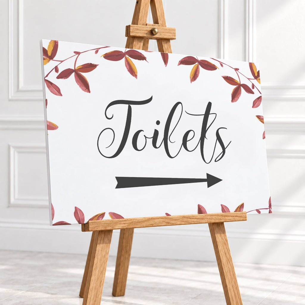 A rectangular white sign with the word "Toilets" in black script and a black right-pointing arrow, surrounded by a border of red and brown leaves. The sign is displayed on a small wooden easel in a bright indoor setting with white paneled walls.