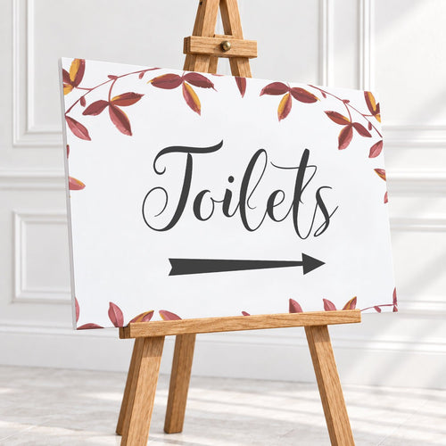 A rectangular white sign with the word "Toilets" in black script and a black right-pointing arrow, surrounded by a border of red and brown leaves. The sign is displayed on a small wooden easel in a bright indoor setting with white paneled walls.
