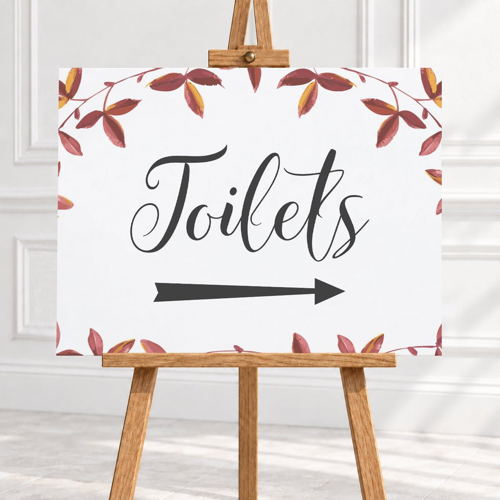 A rectangular white sign with the word "Toilets" in large black script, a black right-pointing arrow below, and a border of red and brown leafy vines, displayed on a wooden easel against a light interior wall.
