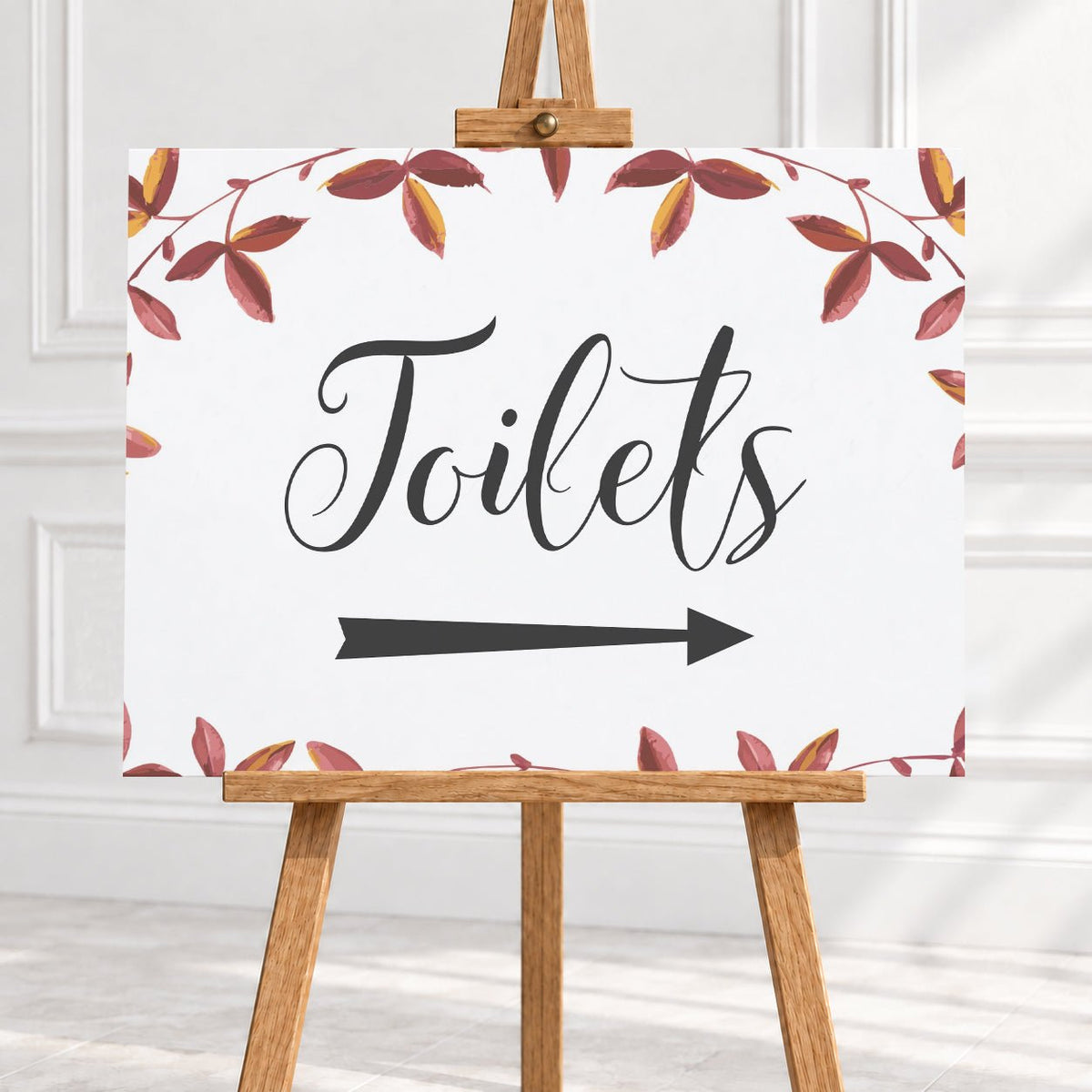 A rectangular white sign with the word "Toilets" in large black script, a black right-pointing arrow below, and a border of red and brown leafy vines, displayed on a wooden easel against a light interior wall.