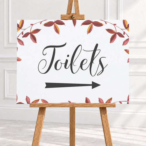 A rectangular white sign with the word "Toilets" in large black script, a black right-pointing arrow below, and a border of red and brown leafy vines, displayed on a wooden easel against a light interior wall.