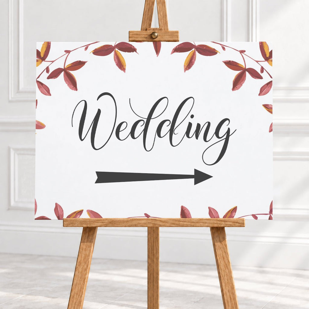 A wedding sign on a wooden easel, featuring the word 'Wedding' in black script with a directional arrow pointing right and a border of watercolor autumn leaves in red, brown, and yellow on a white background.