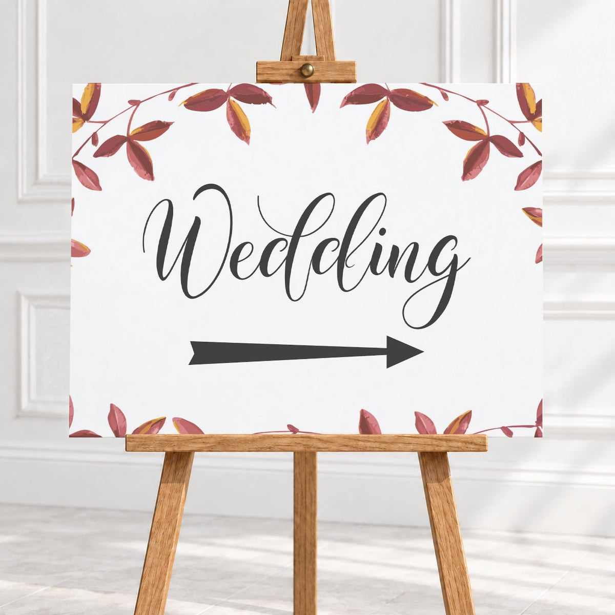 A wedding sign on a wooden easel, featuring the word 'Wedding' in black script with a directional arrow pointing right and a border of watercolor autumn leaves in red, brown, and yellow on a white background.