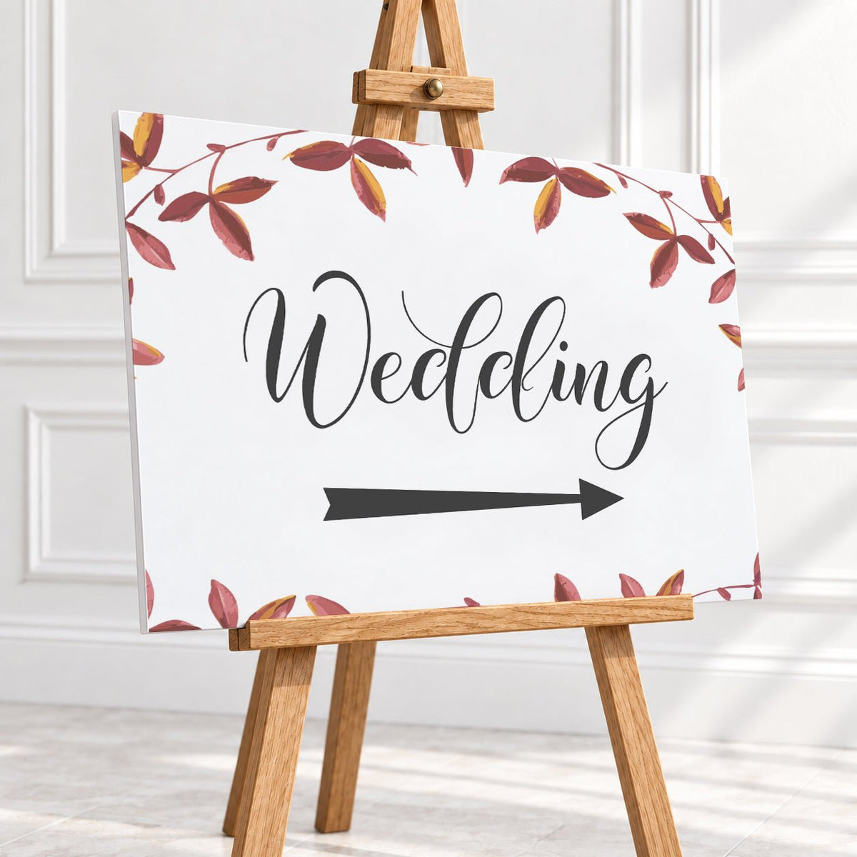 A rectangular wedding sign on a wooden easel, featuring the word 'Wedding' in black script, a black right-pointing arrow, and a border of red and brown leaves on a white background.