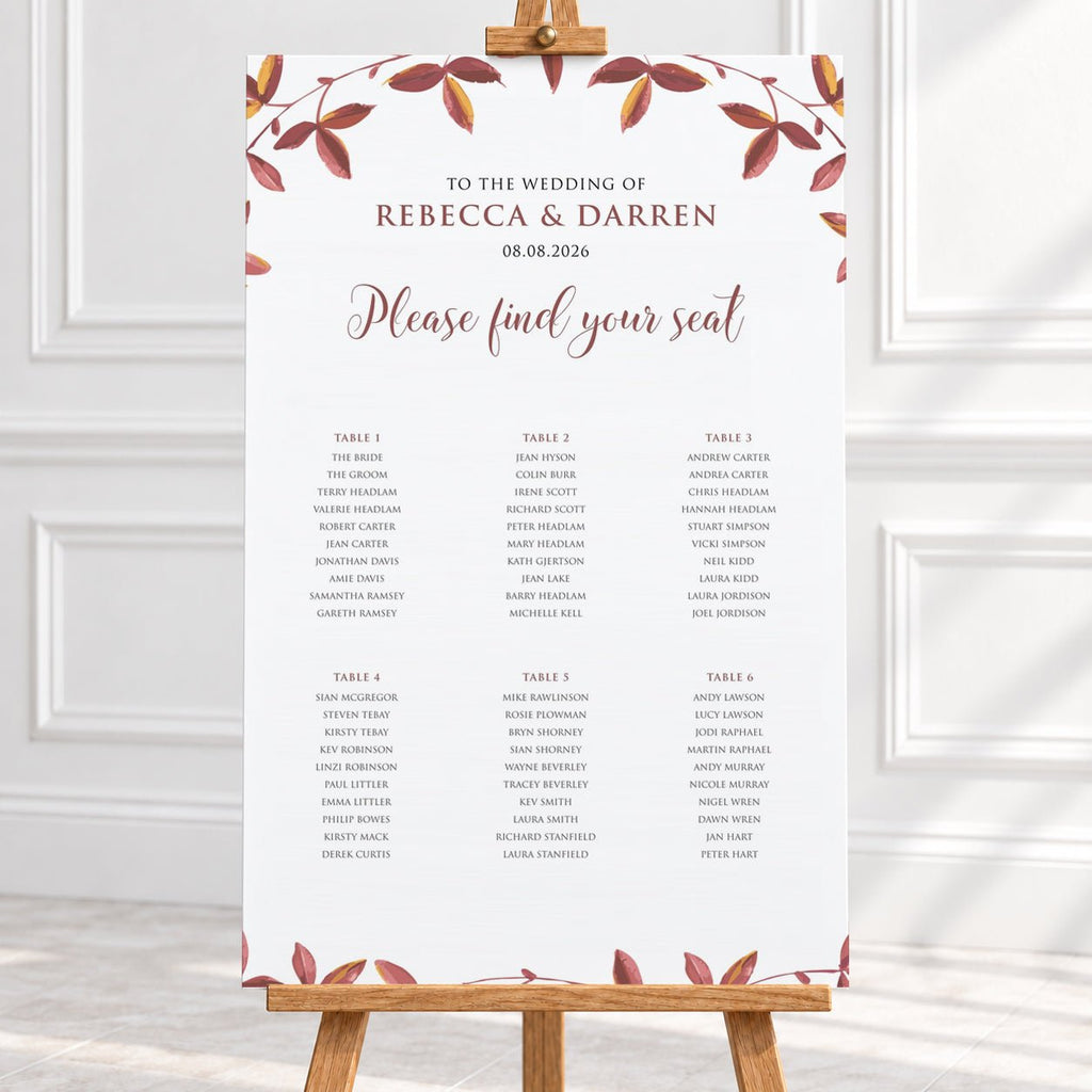 A wedding seating chart print with a white background, elegant brown and dusty rose typography, and a border of autumn leaves in muted red, brown, and gold tones with 6 wedding tables. The print is shown hanging from a wooden hanger on a light-colored wall above a white surface.