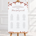A wedding seating chart print with a white background, elegant brown and dusty rose typography, and a border of autumn leaves in muted red, brown, and gold tones with 6 wedding tables. The print is shown hanging from a wooden hanger on a light-colored wall above a white surface.