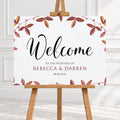 A personalized wedding welcome sign with a watercolor autumn leaf border, displayed on a wooden easel. The sign features the text 'Welcome to the wedding of Rebecca & Darren' and the date '08.