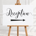 A white reception sign with black script lettering and a black arrow pointing right, displayed on a wooden easel in a bright indoor setting.