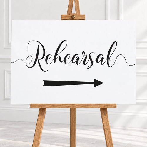 A white sign with black script reading 'Rehearsal' and a black arrow pointing right, displayed on a wooden easel in a bright indoor setting.