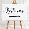 A white rectangular sign with black script reading "Restrooms" and a black arrow pointing right, displayed on a wooden easel in a bright indoor room.