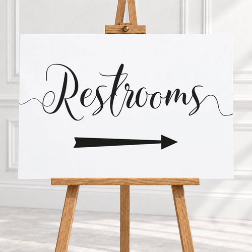 A white rectangular sign with black script reading "Restrooms" and a black arrow pointing right, displayed on a wooden easel in a bright indoor room.