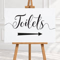 A white rectangular sign with black script reading 'Toilets' and a black arrow pointing right, displayed on a wooden easel in a bright indoor setting.