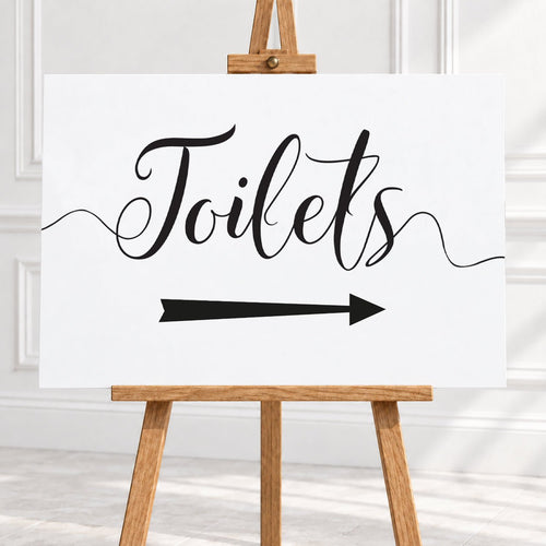 A white rectangular sign with black script reading 'Toilets' and a black arrow pointing right, displayed on a wooden easel in a bright indoor setting.
