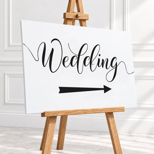 A white wedding sign with black script reading 'Wedding' and a black arrow pointing right, displayed on a wooden easel in a bright white interior setting.