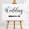 A white wedding sign with black script reading 'Wedding' and a black directional arrow, displayed on a wooden easel in a bright indoor setting.