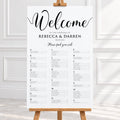 A wedding welcome sign printed on a white sheet, displayed on a wooden easel in a bright indoor setting with white paneled walls.