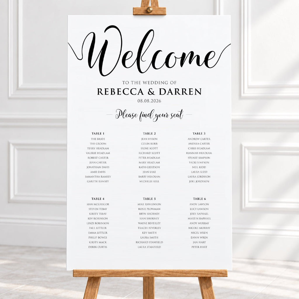 A wedding welcome sign displayed on a wooden easel, featuring elegant black script and serif text on a white background, set against a bright, softly lit interior backdrop.