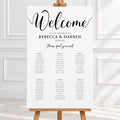 A wedding welcome sign displayed on a wooden easel, featuring elegant black script and serif text on a white background, set against a bright, softly lit interior backdrop.