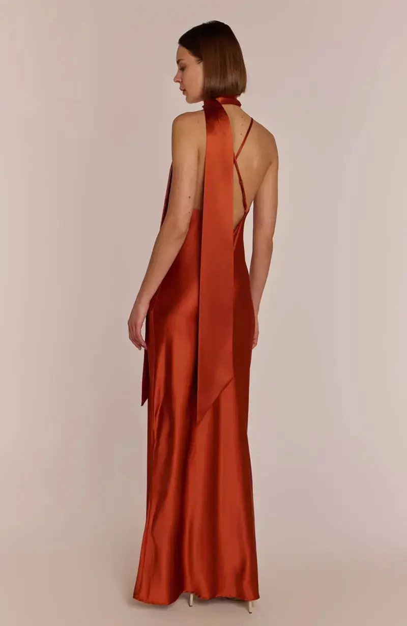 Bridesmaid wearing a rust-colored burnt orange halter neck dress with a draped front on a plain background