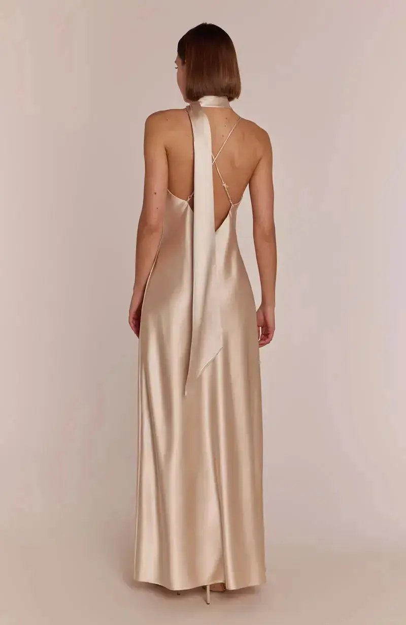 Woman wearing a long, elegant champagne Bridesmaid dress with a backless design on a plain background