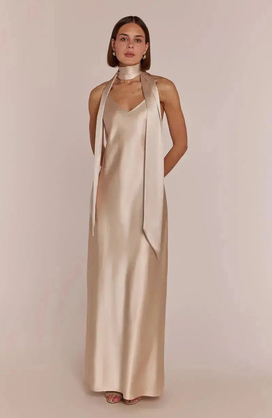 Bridesmaid wearing a long, champagne satin dress with thin straps on a plain background