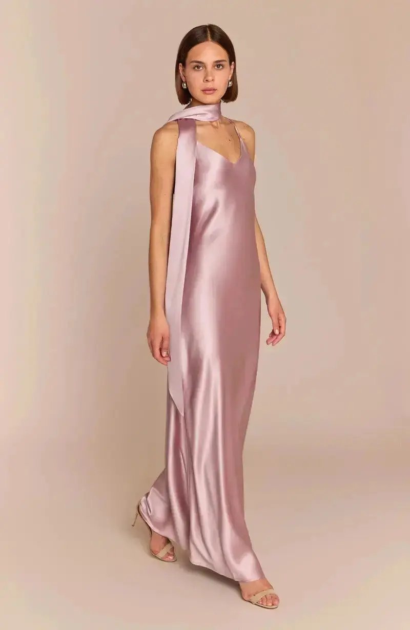 A woman models the Brooklyn bridesmaid Dress in Rose Pink Satin, featuring thin straps and a matching long scarf draped around her neck. Her shoulder-length brown hair stands out against a plain beige background.
