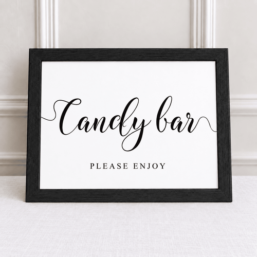 A black-framed sign with the words 'Candy bar' in black script and 'PLEASE ENJOY' below in a serif font, displayed against a light neutral wall and tabletop.