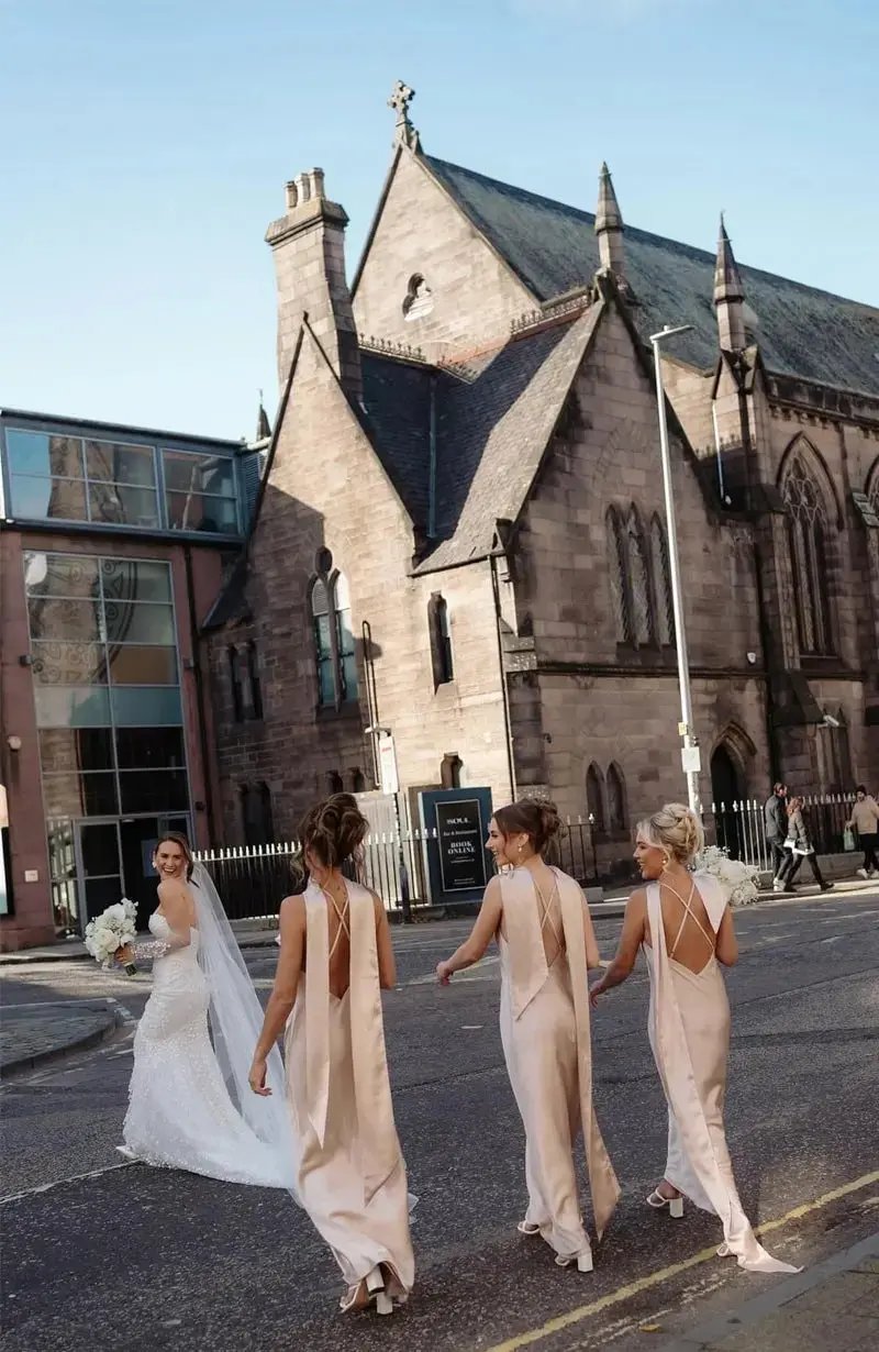 Three bridesmaids in elegant champagne bridesmaid dresses walking behind the bride on a street with a church in the background