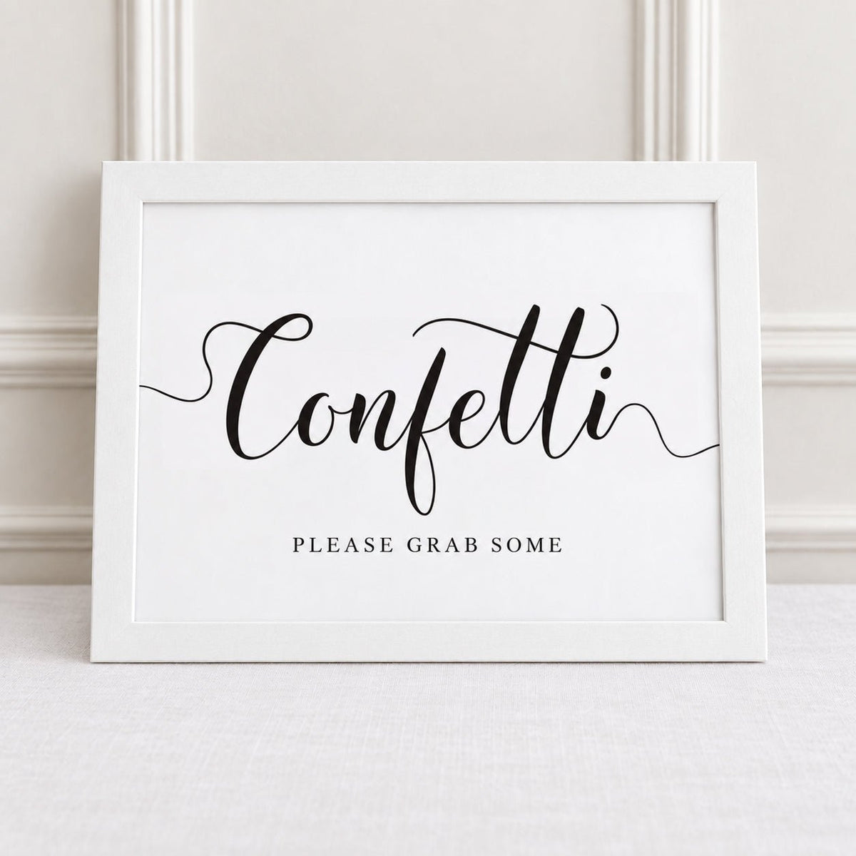 A white framed sign with black script reading 'Confetti' and smaller text 'PLEASE GRAB SOME' below, leaning against a light wall on a pale surface.