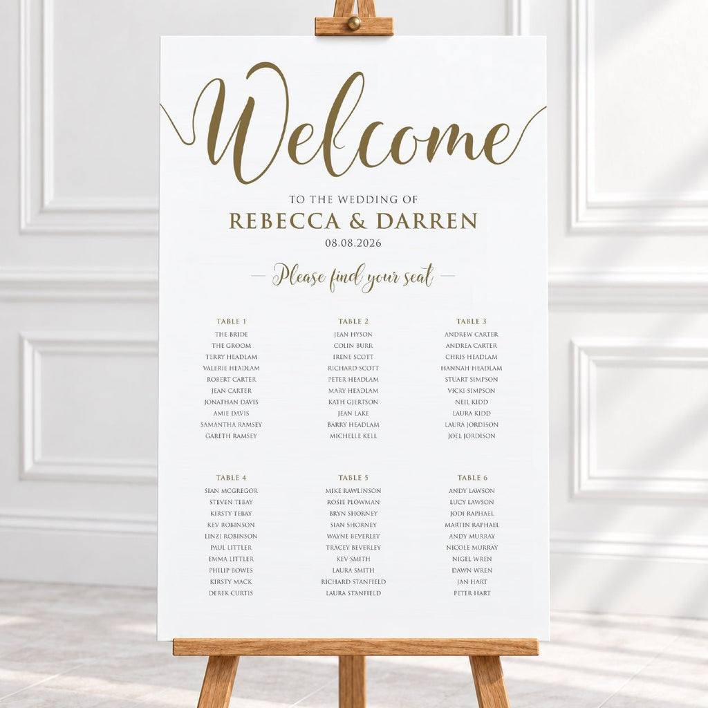 A wedding welcome sign printed on a white board with elegant gold script and black table seating details, displayed on a wooden easel in a bright indoor setting.